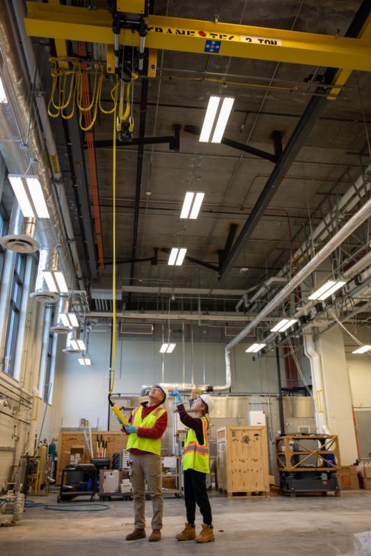 A man and woman in yellow vests are hard hats in a lab with a yellow crane.